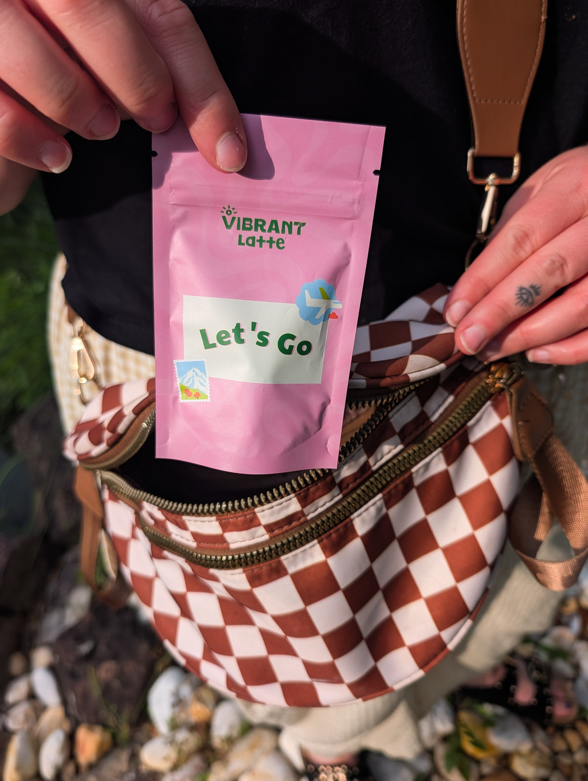 A person outdoors holds up a pink vibrant Latte 'Let's Go' pouch next to a brown and white checkered crossbody bag.