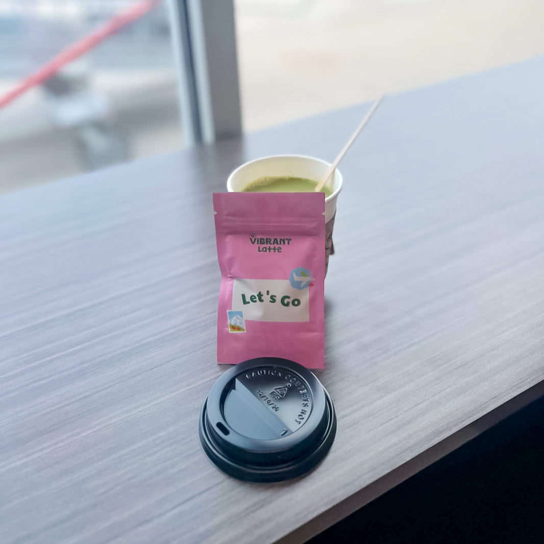 A pink Vibrant Latte 'Let's Go' matcha pouch stands on a light gray wooden table next to a paper cup of green matcha with a stirrer and a black lid, in an airport