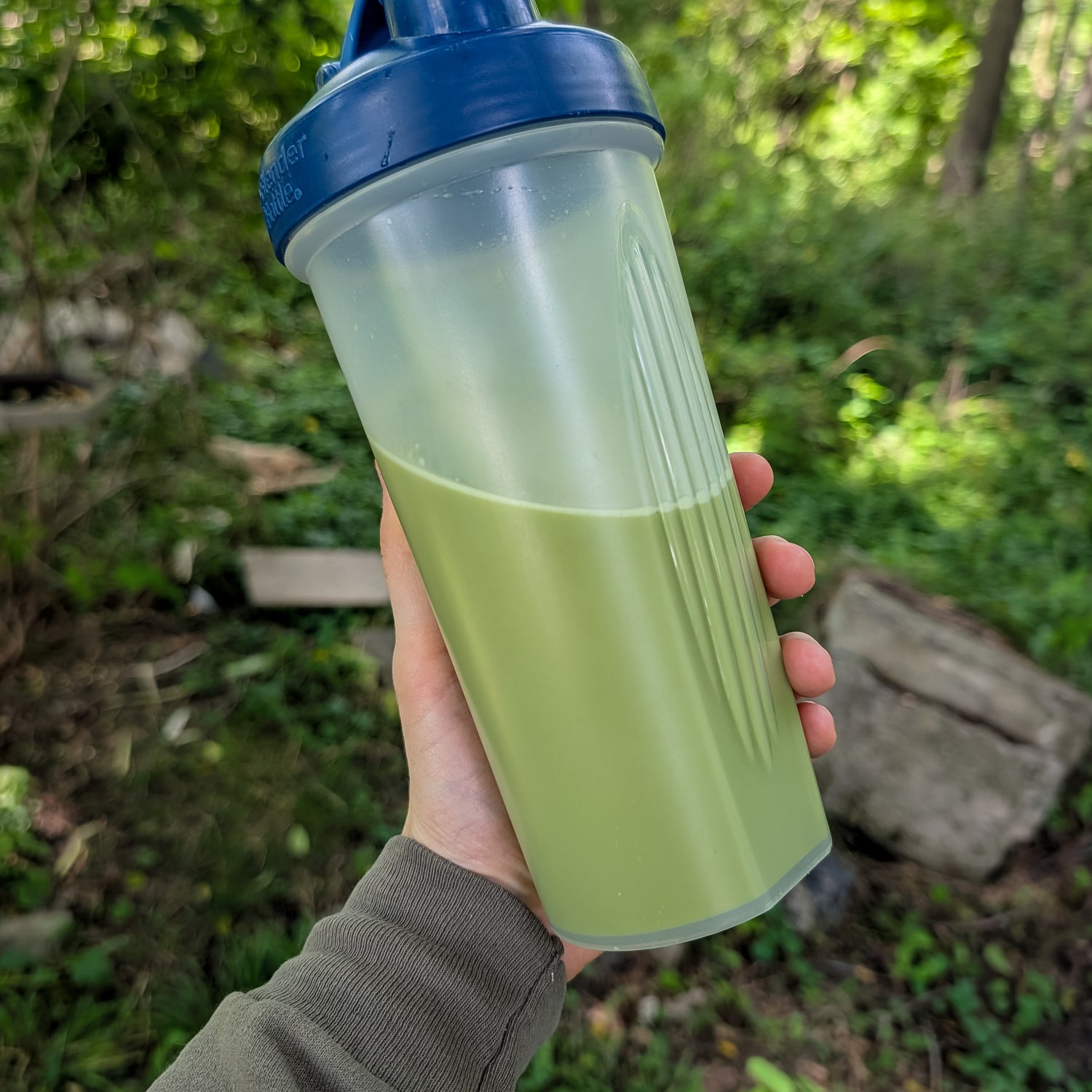 Hand holding instant matcha latte in a clear shaker bottle with a blue lid against a natural background.