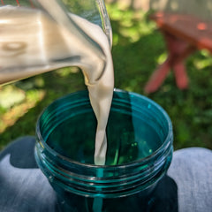 A close-up action shot of a creamy instant oatmilk being poured from a clear glass pitcher into a translucent dark teal plastic tumbler, held outdoors on a person's lap with a blurred green grassy background.