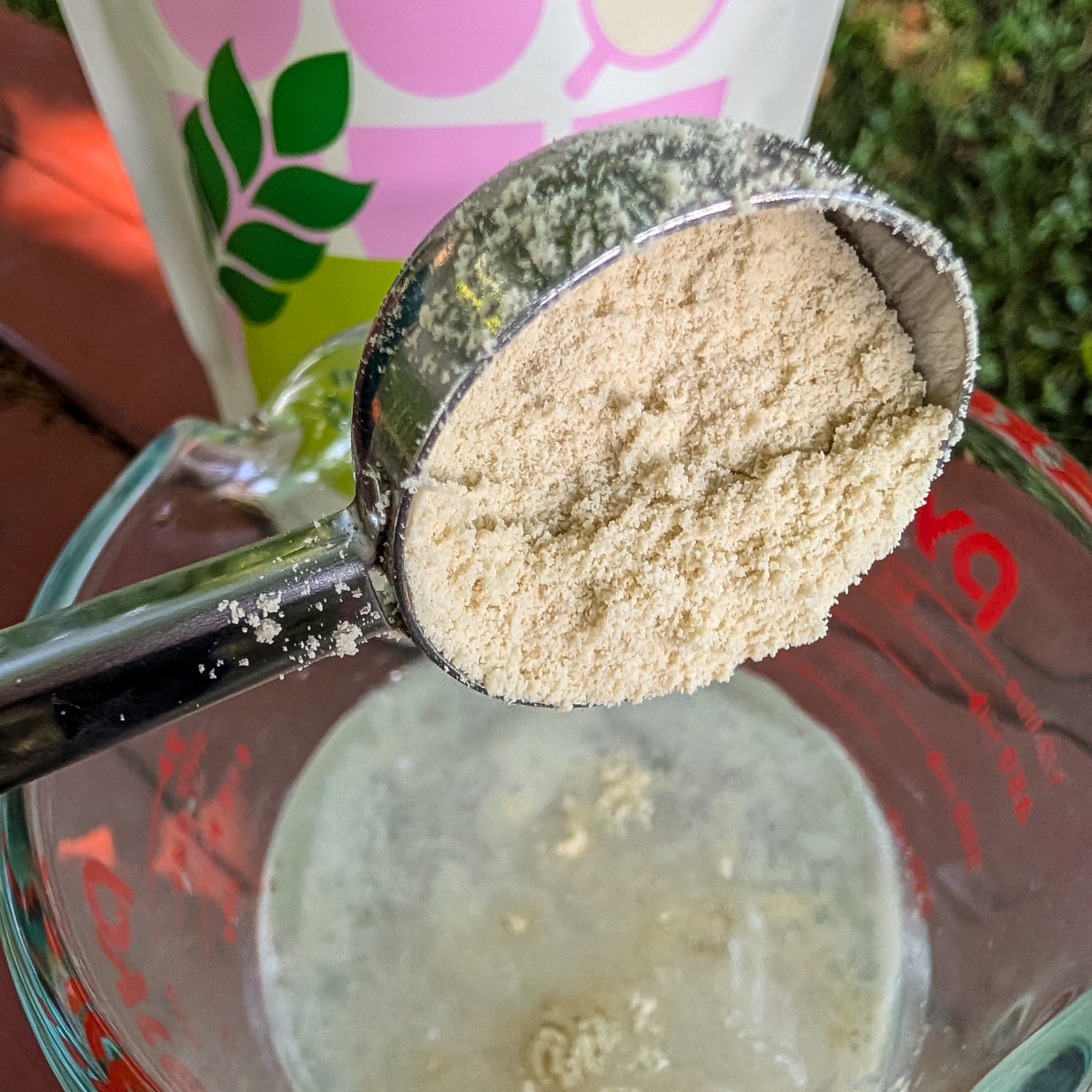 A metal measuring scoop filled with instant oatmilk powder, held above a glass measuring cup containing a pale liquid, with a branded bag blurred in the background.