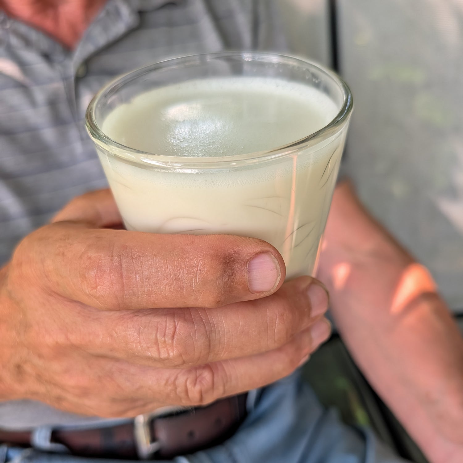 A person holding a clear glass tumbler filled with oatmilk, photographed closely with the hand and torso visible against an outdoor background.