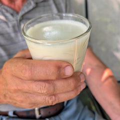 A person holding a clear glass tumbler filled with oatmilk, photographed closely with the hand and torso visible against an outdoor background.