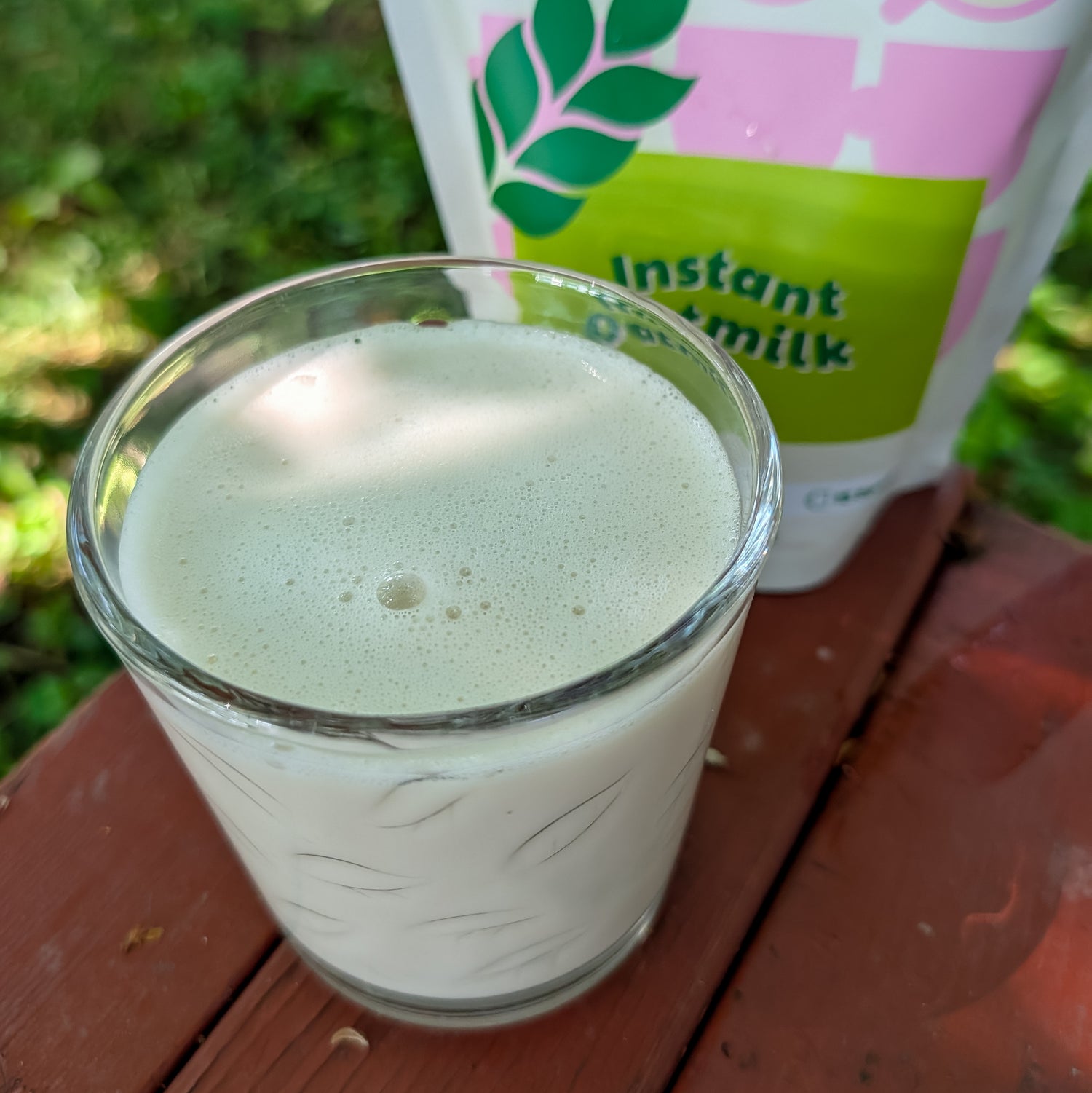 A glass of frothy instant oatmeal milk sits on a wooden table outdoors, with a pouch of instant oatmilk in the background.