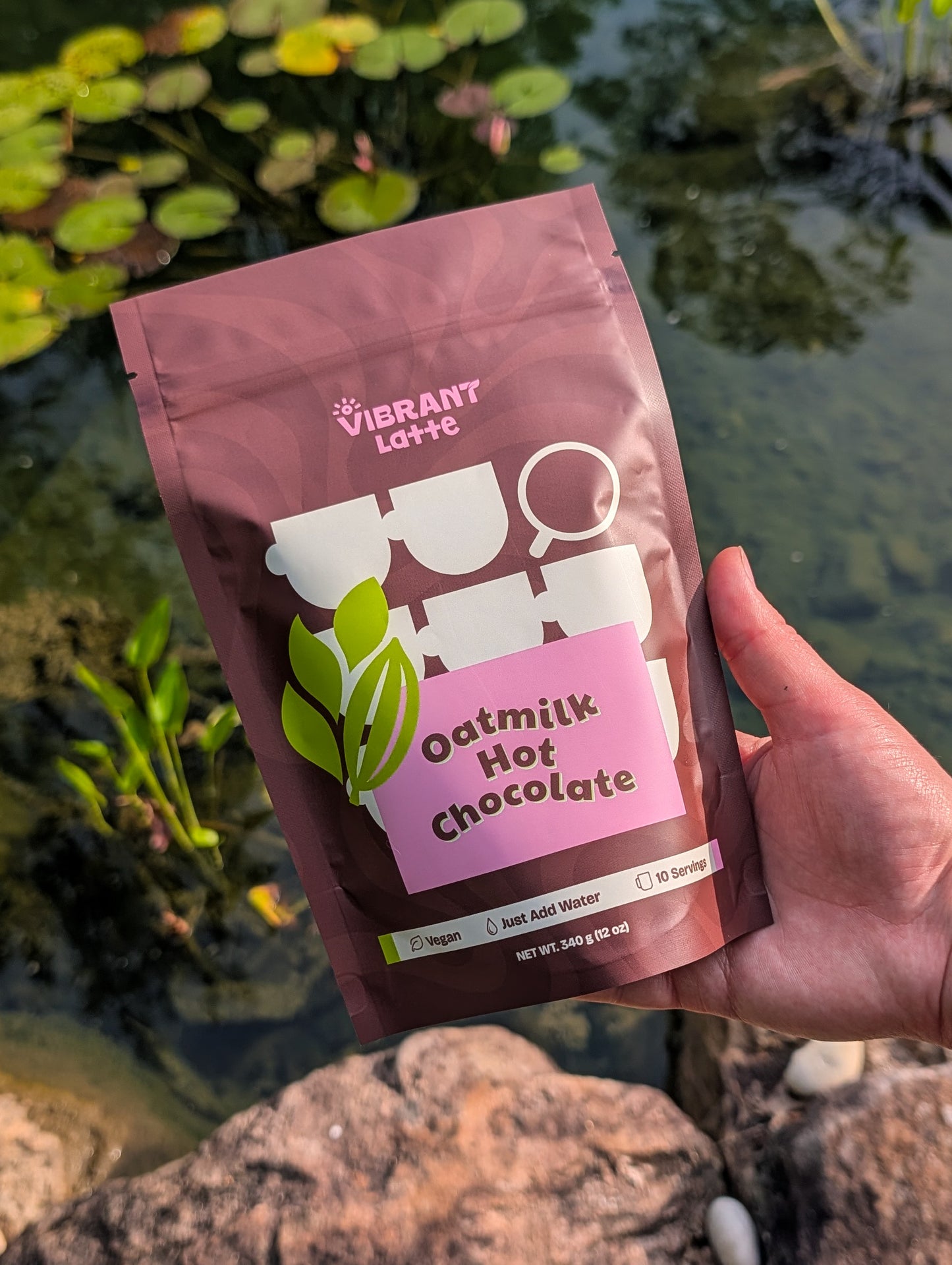 Hand holding a package of Vibrant Latte Oatmilk Hot Chocolate in front of a natural background with water and plants.