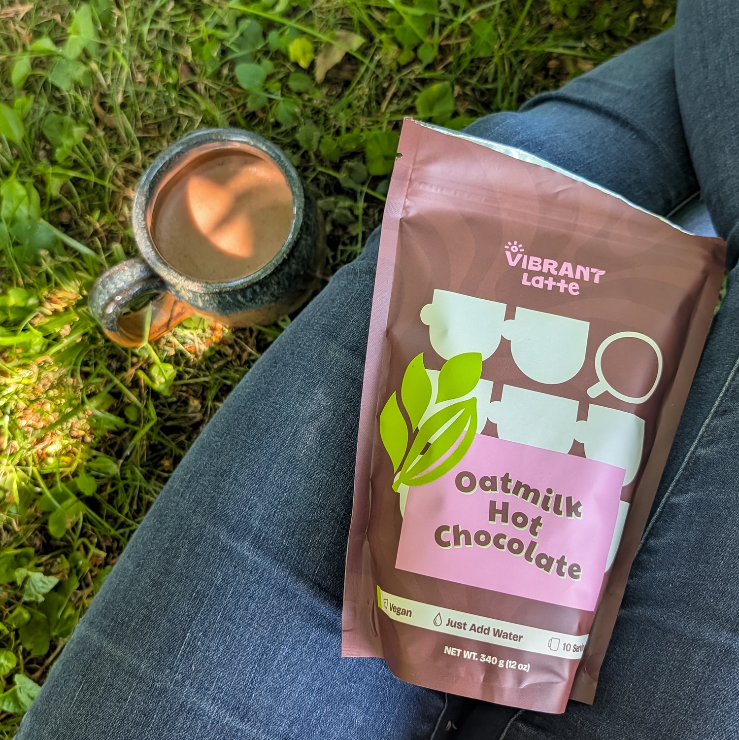 A bag of Vibrant Latte Oatmilk Hot Chocolate sits on a person's lap outdoors, next to a ceramic mug filled with hot chocolate on grass.