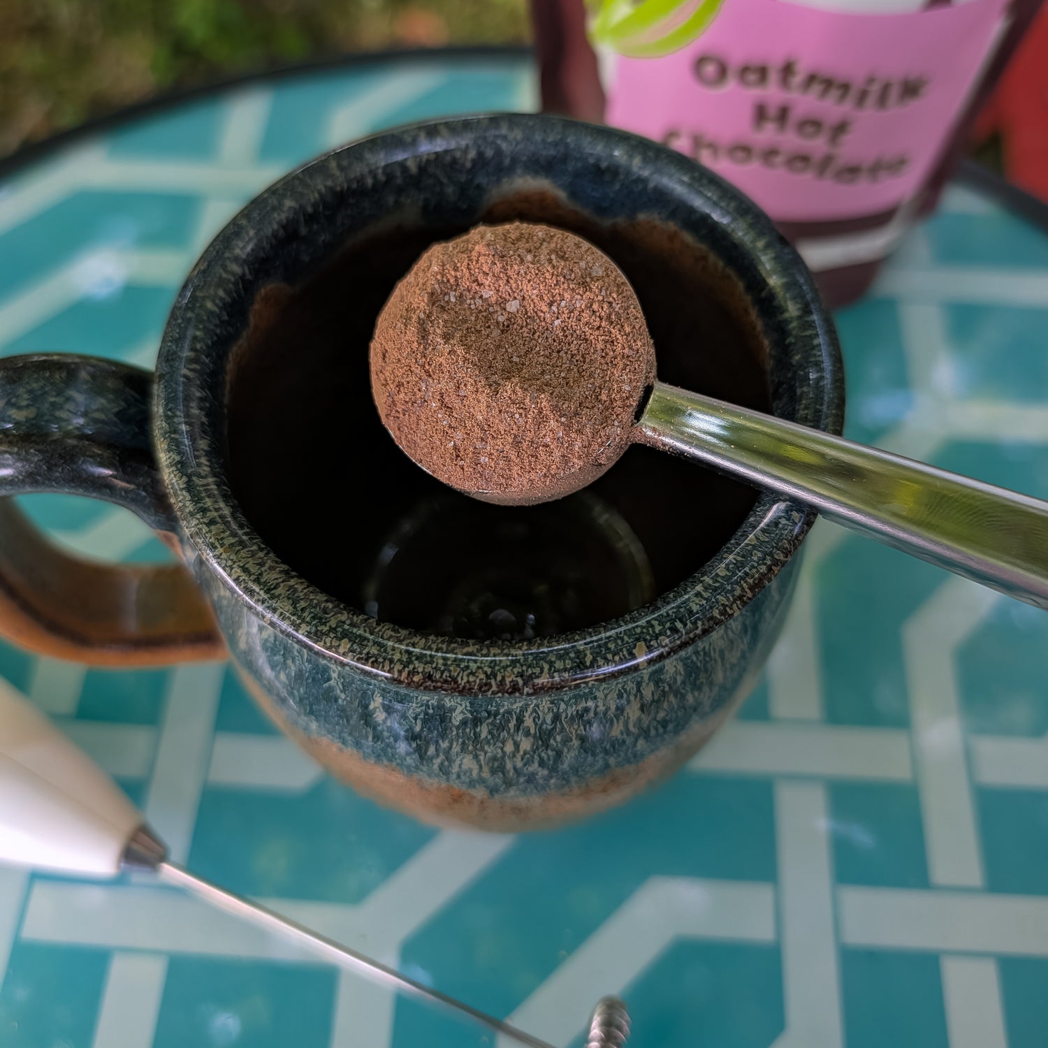 A close-up of a ceramic mug filled with hot chocolate, with a spoon holding a scoop of hot chocolate powder above the mug. A bag labeled 'Oatmilk Hot Chocolate' is blurred in the background on a teal patterned table.