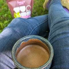 A close-up of a ceramic mug filled with hot chocolate resting on a person's lap, with blue jeans visible and a pink bag of instant oatmilk in the blurred background on grass.