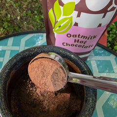 A close-up of a mug with a metal scoop holding hot chocolate being added. A pink and brown bag labeled 'Oatmilk Hot Chocolate' stands behind the bowl on a teal patterned table outdoors.