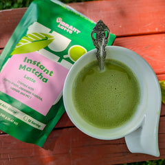 Green matcha latte in a white cup with a spoon, next to a Vibrant Latte instant matcha latte package on a wooden surface