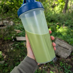 Hand holding instant matcha latte in a clear shaker bottle with a blue lid against a natural background.