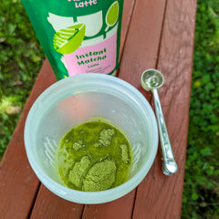 Instant matcha latte powder in a clear container with a spoon and matcha packaging on a wooden surface