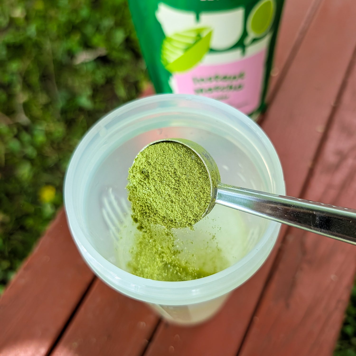 Spoonful of green instant matcha latte poweder over a clear container on a wooden surface with a blurred background